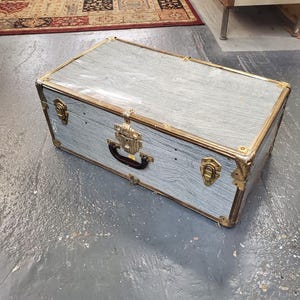 Puede incluir: Una maleta vintage de color gris con detalles dorados y un asa negra. La maleta tiene un patrón de veta de madera y está en buen estado.