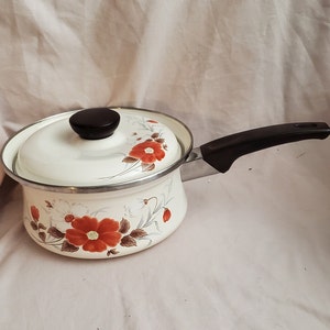 Pode incluir: Caçarola de esmalte branco com alça preta e design floral com flores vermelhas e brancas. A tampa tem uma pega preta.