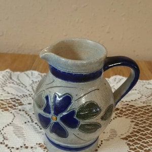 Op de afbeelding: Een kleine, grijze keramische kan met een blauwe handgreep en een bloemenmotief. De kan heeft een blauwe bloem met een gele kern en groene bladeren.