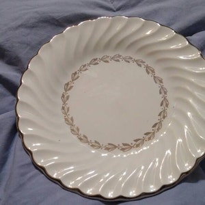 May include: A white ceramic plate with a gold rim and a decorative leaf pattern around the center. The plate has a swirled design around the edge.
