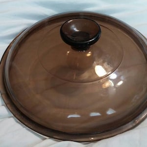 Puede incluir: Una tapa de vidrio marrón con un pomo en la parte superior. La tapa es redonda y tiene una superficie lisa.