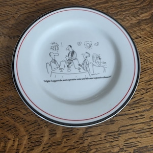 May include: White ceramic plate with a black and red rim, featuring a cartoon illustration of people at a table. The plate has the text "Might I suggest the most expensive wine and the most expensive dinner?"