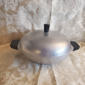 Peut inclure: Un plat en aluminium argenté avec un couvercle et des poignées noirs. Le plat a une surface texturée.