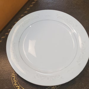 Może przedstawiać: Biały porcelanowy talerz obiadowy z delikatnym wzorem kwiatowym wokół krawędzi. Talerz ma złotą obwódkę.