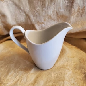 May include: A white ceramic creamer with a curved handle. The creamer is sitting on a cream-coloured fabric with a floral pattern.