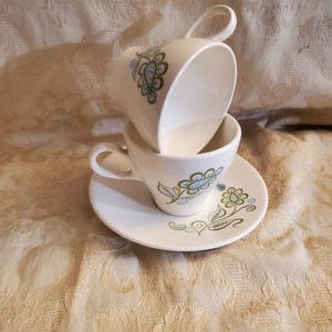 May include: Two white teacups with a floral design in shades of green and blue. The cups are stacked on top of each other and are sitting on a white saucer with a matching floral design.