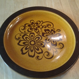 Puede incluir: Un plato de cena de cerámica marrón y negro con un diseño floral. El plato tiene un borde marrón y un patrón floral marrón y negro en el centro.