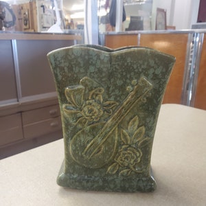 Puede incluir: Un jarrón de cerámica verde con un diseño en relieve de una guitarra y flores. El jarrón tiene un esmalte moteado.