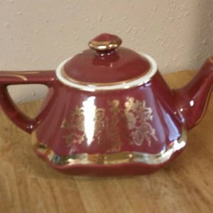 May include: A burgundy teapot with a white rim and gold floral design. The teapot has a rounded body and a spout on the side.