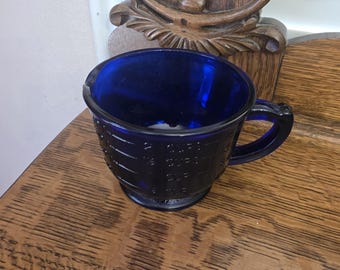 May include: A cobalt blue glass measuring cup with a handle. The cup has measurement markings for cups and ounces. The cup is sitting on a wooden surface.