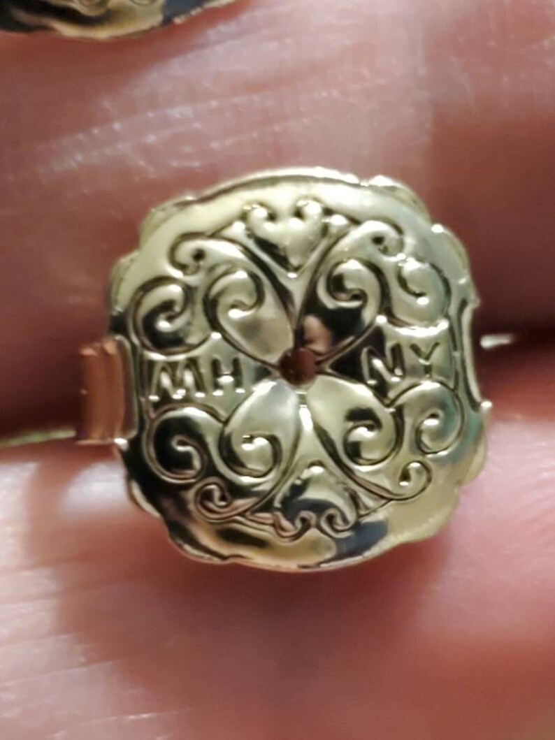 May include: Gold-toned metal ring with an ornate design featuring a central circle and the letters "MH" and "NY" on either side.