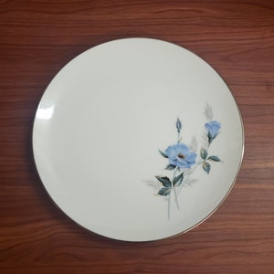 Puede incluir: Un plato de cena de cerámica blanca con un borde dorado y un diseño floral azul. Las flores son estilizadas y tienen un aspecto delicado pintado a mano.