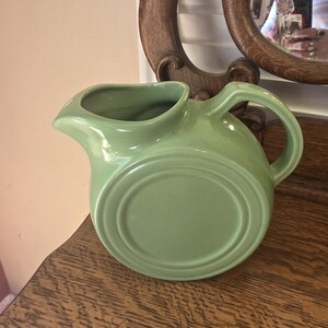 May include: A light green ceramic pitcher with a rounded body, a spout, and a handle. The pitcher has a circular base and a smooth, glossy finish. The pitcher is sitting on a wooden surface.