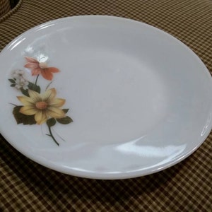 May include: A white ceramic plate with a floral design featuring yellow, orange, and white flowers. The plate has a slightly raised rim.