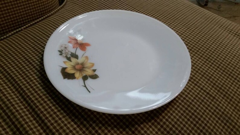 May include: A white ceramic plate with a floral design featuring yellow, orange, and white flowers. The plate has a slightly raised rim.