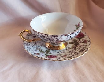 May include: A white teacup and saucer set with a floral pattern in shades of pink, purple, and green. The set has a gold trim and a delicate design.