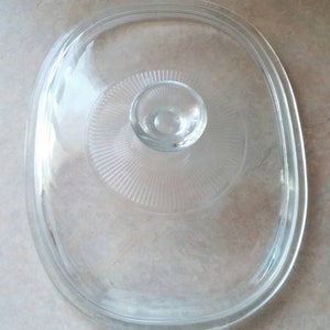 May include: A clear glass lid for a casserole dish. The lid has a knob in the center and a raised rim around the edge.