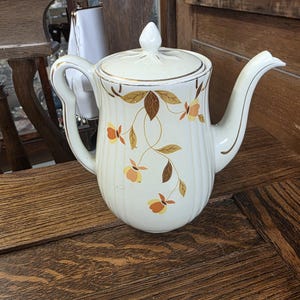 Puede incluir: Tetera de cerámica color crema con tapa, adornada con ribetes dorados y motivos florales en tonos marrón, naranja y amarillo. La tetera tiene un pico y asa curvos, colocada sobre una superficie de madera.