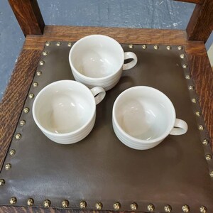 以下が含まれることがあります： 茶色の革張りの座席を背景に、白いセラミック製のマグカップが3つあります