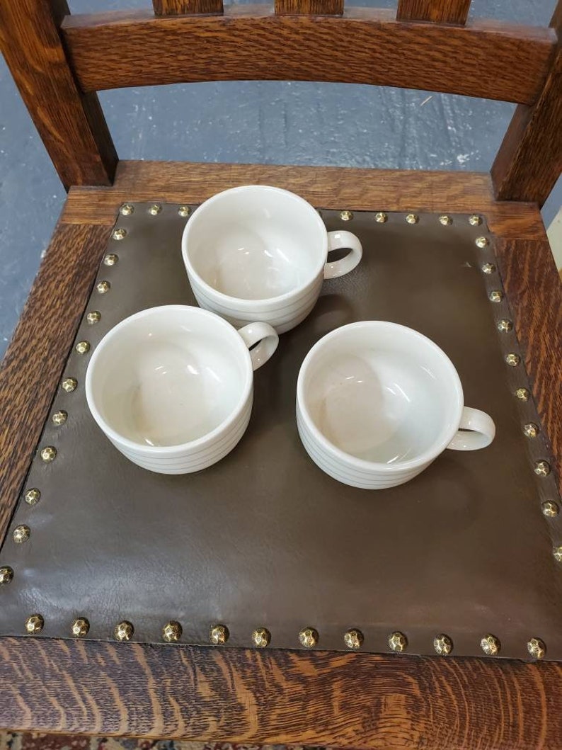 以下が含まれることがあります： 茶色の革張りの座席を背景に、白いセラミック製のマグカップが3つあります