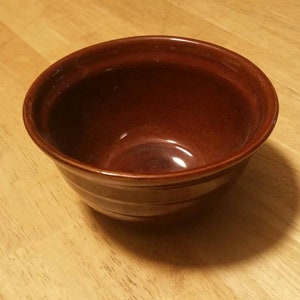 May include: A brown ceramic bowl with a smooth, glossy finish. The bowl is empty and has a slight curve to the sides.