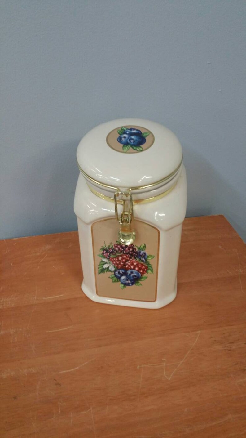 K&ouml;nnte beinhalten: Ein wei&szlig;er Keramiktopf mit Deckel und goldfarbenem Verschluss. Der Topf hat ein dekoratives Etikett mit einem Blaubeer- und Beeren-Design. Der Deckel hat auch ein Blaubeer-Design. Der Topf steht auf einer Holzoberfl&auml;che.