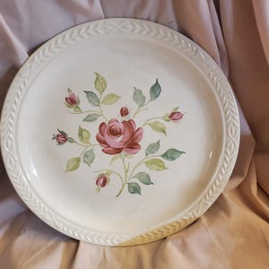 Könnte beinhalten: Ein weißer Keramikteller mit einem erhabenen Randdesign. Der Teller zeigt ein handgemaltes Blumenmuster mit einer rosa Rose und grünen Blättern.