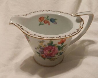 May include: A white porcelain creamer with a gold trim and floral design. The creamer has a handle and a spout.