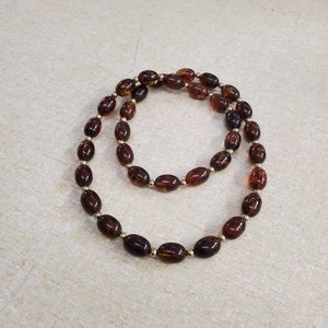 Puede incluir: Un collar con cuentas ovaladas de color ámbar oscuro, ensartadas con pequeñas cuentas doradas. El collar se exhibe sobre una superficie neutra, resaltando su rico color y diseño. Una pieza de joyería.