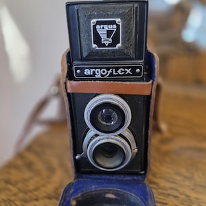 May include: A vintage black Argus Argoflex camera with a brown leather strap and a blue velvet interior. The camera has two lenses and the name "argoflex" is printed on the front.