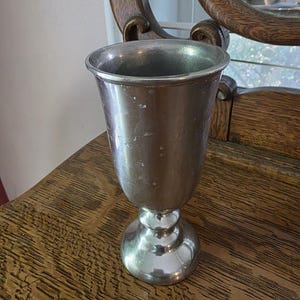 May include: A silver-colored metal goblet with a wide rim and a rounded base. The goblet has a classic shape and is sitting on a wooden surface. The goblet is likely vintage or antique.