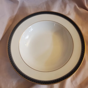 Puede incluir: Un tazón de sopa de cerámica blanca con un borde negro y dorado. El borde tiene un patrón de cuadros.