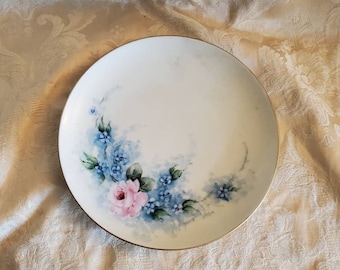 May include: A white porcelain plate with a gold rim features a hand-painted design of a pink rose and blue forget-me-not flowers.