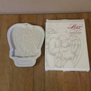 Puede incluir: Un cortador de galletas de cerámica blanca con forma de ángel que sostiene una cesta de flores. El cortador está en su caja original, que también es blanca y presenta el texto "Love The 1990 Langsberger Pottery Angel Series Cookie Mold".