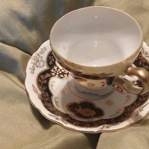 May include: A white teacup and saucer with a black and gold floral design. The cup has a gold rim and a gold handle. The saucer has a scalloped edge.