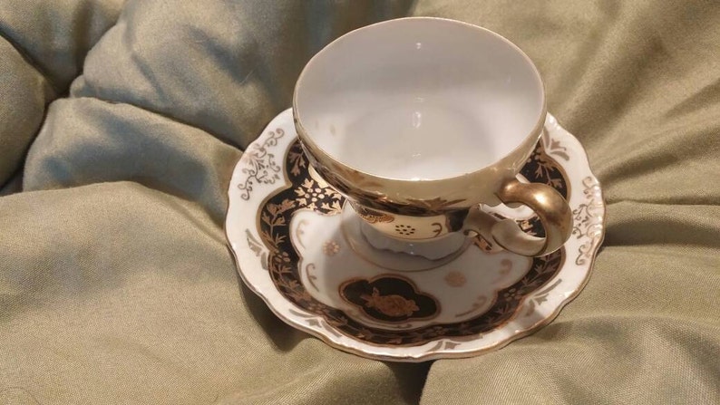 May include: A white teacup and saucer with a black and gold floral design. The cup has a gold rim and a gold handle. The saucer has a scalloped edge.