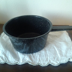 Peut inclure: Une casserole en émail noir avec une finition mouchetée et une poignée noire. La casserole est posée sur un torchon blanc à bord festonné.