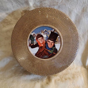 May include: A gold-rimmed ceramic plate with a woven pattern. The center of the plate features a vintage Christmas scene with a man and a child in a snowy town.
