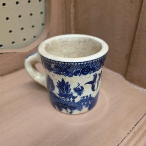 May include: Small, off-white ceramic mug with a blue Willow pattern design. The mug has a small handle and a cylindrical shape. The blue design features a landscape with buildings, trees, and birds. The mug is likely vintage.