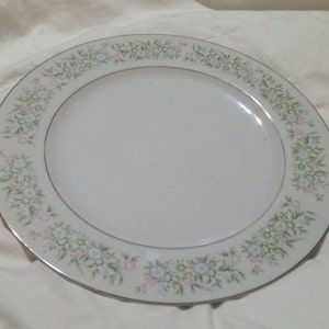 May include: A white ceramic dinner plate with a floral design in shades of green, pink, and white. The plate has a silver rim.