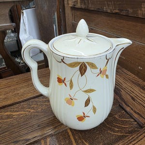 Puede incluir: Jarra de cerámica color crema con tapa. La jarra presenta un diseño floral en tonos marrones, naranjas y amarillos. El asa y el pico están acentuados con ribetes dorados. La tapa tiene un pequeño pomo en la parte superior. La jarra está sobre una superficie de madera.