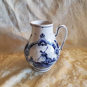 May include: White ceramic pitcher with a blue and white windmill design. The pitcher has a curved handle and a wide spout. The windmill scene is surrounded by floral patterns. The pitcher is likely for serving beverages.