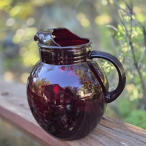 Puede incluir: Una jarra de vidrio rojo rubí vintage con un cuerpo redondeado y un asa curva. La jarra tiene un pico para verter y una tapa. El vidrio refleja el entorno. La jarra está sobre una superficie de madera.