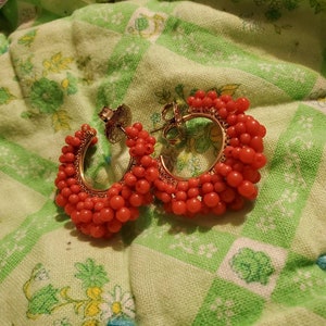May include: A pair of hoop earrings with a gold base and red beads. The beads are clustered together to form a thick hoop.