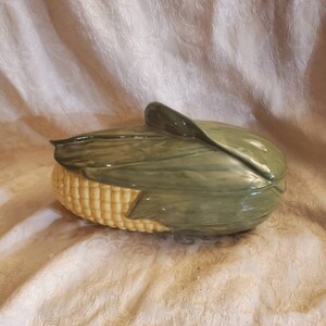 May include: A ceramic corn on the cob serving dish with a green lid. The dish is shaped like an ear of corn with yellow kernels and green husks.
