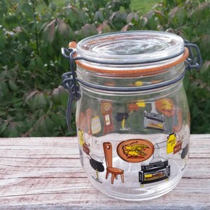 May include: A clear glass jar with a black metal lid and a rubber seal. The jar has a colorful design of various household items, including a chair, a stove, and a clock.