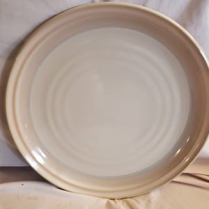 May include: A large, round, white ceramic serving platter with a light brown rim and subtle concentric circles in the centre.