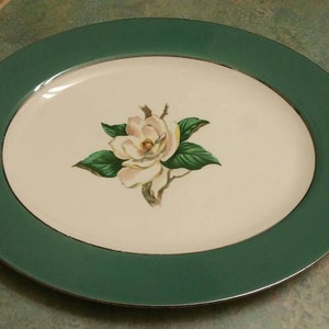 May include: A white oval plate with a green rim features a single white magnolia flower with green leaves in the centre.