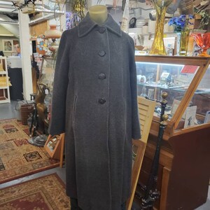 May include: A long, gray wool coat with a collar and five black buttons down the front. The coat is on a mannequin in a shop setting.