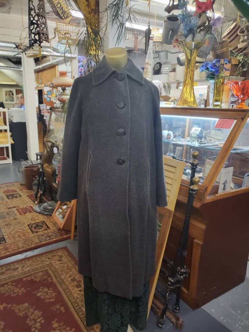 May include: A long, gray wool coat with a collar and five black buttons down the front. The coat is on a mannequin in a shop setting.